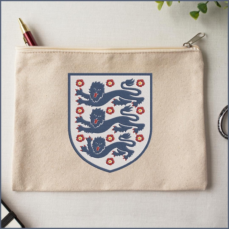 Puede incluir: Un estuche de lona beige con cremallera que presenta un emblema de escudo centrado. El escudo muestra tres leones azul oscuro en una pila vertical, rodeados por peque&ntilde;os iconos de rosas Tudor rojas y amarillas, todo en un borde azul.