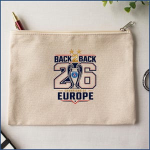 Pode incluir: Uma bolsa de lona bege com fecho de correr, apresentando uma impress&atilde;o gr&aacute;fica que diz BACK 2 BACK CHAMPIONS OF EUROPE com o n&uacute;mero 26, duas estrelas douradas e uma ilustra&ccedil;&atilde;o central de um trof&eacute;u com o log&oacute;tipo do Paris Saint-Germain.