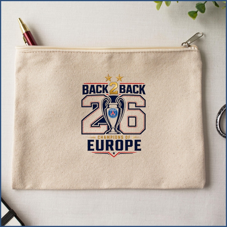 Pode incluir: Uma bolsa de lona bege com fecho de correr, apresentando uma impress&atilde;o gr&aacute;fica que diz BACK 2 BACK CHAMPIONS OF EUROPE com o n&uacute;mero 26, duas estrelas douradas e uma ilustra&ccedil;&atilde;o central de um trof&eacute;u com o log&oacute;tipo do Paris Saint-Germain.