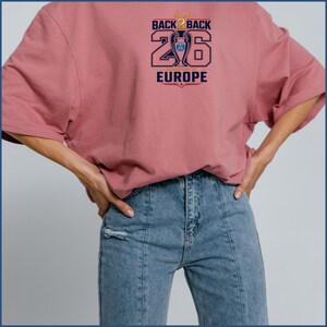 Pode incluir: Uma t-shirt de manga curta em rosa seco com uma impress&atilde;o gr&aacute;fica que diz BACK 2 BACK 26 EUROPE, um &iacute;cone de trof&eacute;u central e duas estrelas douradas. O design &eacute; impresso em azul-marinho, dourado e vermelho no peito da pe&ccedil;a.
