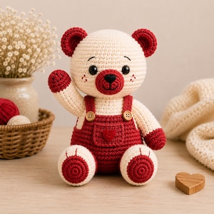 Peut inclure: Un ours en peluche crocheté de couleur crème avec des oreilles, des pattes et un museau rouge foncé. L'ours porte une salopette crochetée rouge foncé avec deux petits boutons en bois sur les bretelles et un petit cœur rouge sur la poche.