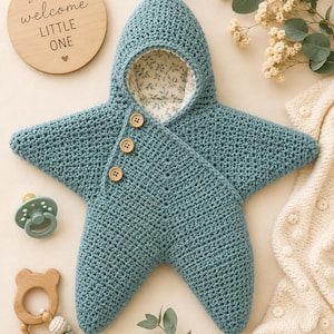 Peut inclure: Une gigoteuse en forme d'étoile crochetée bleu sarcelle avec une capuche surmontée d'un petit pompon. Le vêtement comporte trois boutons en bois brun clair sur le devant et est doublé d'un tissu blanc imprimé d'un motif de feuilles vertes.