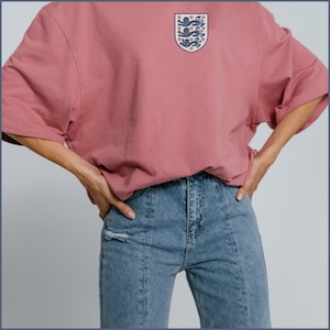 Puede incluir: Una camiseta oversize de color rosa empolvado con un peque&ntilde;o emblema en forma de escudo en el pecho izquierdo. El emblema muestra tres leones azules apilados verticalmente sobre un fondo blanco con detalles rojos. Se combina con vaqueros azules.