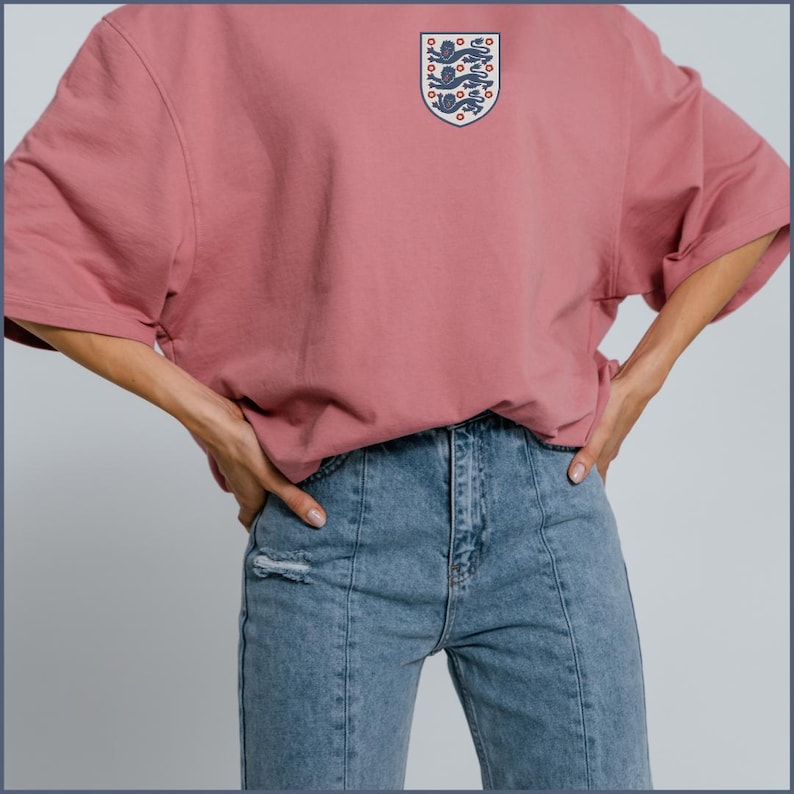 Puede incluir: Una camiseta oversize de color rosa empolvado con un peque&ntilde;o emblema en forma de escudo en el pecho izquierdo. El emblema muestra tres leones azules apilados verticalmente sobre un fondo blanco con detalles rojos. Se combina con vaqueros azules.