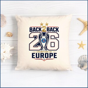 Pode incluir: Uma almofada quadrada em tom branco sujo com uma impress&atilde;o gr&aacute;fica contendo o texto BACK 2 BACK, o n&uacute;mero 26 e CHAMPIONS OF EUROPE. O design inclui um &iacute;cone de trof&eacute;u central com o log&oacute;tipo do Paris Saint-Germain, duas estrelas douradas e detalhes vermelhos.