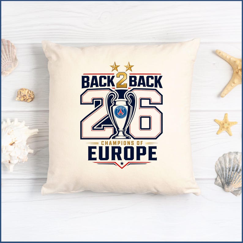 Pode incluir: Uma almofada quadrada em tom branco sujo com uma impress&atilde;o gr&aacute;fica contendo o texto BACK 2 BACK, o n&uacute;mero 26 e CHAMPIONS OF EUROPE. O design inclui um &iacute;cone de trof&eacute;u central com o log&oacute;tipo do Paris Saint-Germain, duas estrelas douradas e detalhes vermelhos.