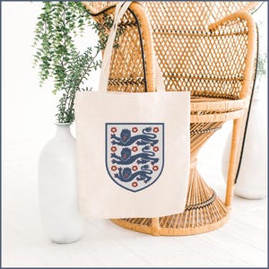 Puede incluir: Una bolsa de tela color crema con un gr&aacute;fico central de un escudo que contiene tres leones azul oscuro apilados verticalmente, intercalados con peque&ntilde;as flores rojas, todo encerrado dentro de un borde azul oscuro en la parte frontal.