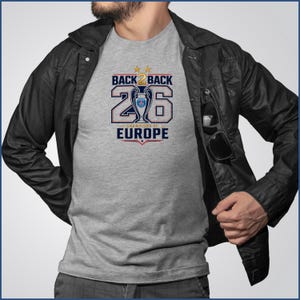 Pode incluir: Uma t-shirt cinzenta mesclada com uma estampa gr&aacute;fica contendo o texto BACK 2 BACK e CHAMPIONS OF EUROPE em azul-marinho e vermelho. O design inclui o n&uacute;mero 26, duas estrelas douradas e uma ilustra&ccedil;&atilde;o central de trof&eacute;u com um log&oacute;tipo azul e vermelho.
