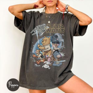 May include: Dark gray t-shirt featuring a mashup of Star Wars and Winnie the Pooh characters. The design includes the Millennium Falcon, the Death Star, and various characters like Pooh, Tigger, and Eeyore. The words "STAR WARS" are printed above the characters.