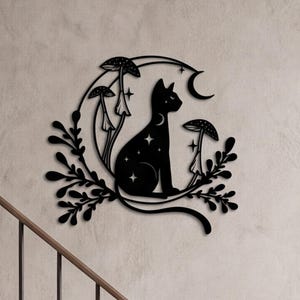 May include: A black metal wall art piece featuring a silhouette of a cat sitting within a crescent moon shape. The design includes three mushrooms, small star cutouts, and leafy vine accents extending from the base of the moon and cat figure.