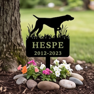 May include: A black metal garden stake featuring a silhouette of a dog standing in grass above a rectangular plaque. The plaque displays the text HESPE and the years 2012-2023 in a light-colored font, centered on the metal surface.