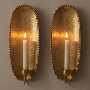 May include: Two oval wall sconces made of hammered brass with a beaded edge. Each sconce features a central candle holder with a single white taper candle. The brass surface has a reflective, metallic finish that catches the light from the flames.
