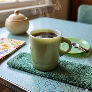 May include: A light green jadeite mug filled with steaming dark coffee. The mug features a smooth, glossy finish and a sturdy handle. It sits on a textured dark green rectangular cloth, showcasing a classic, simple design for hot beverages.