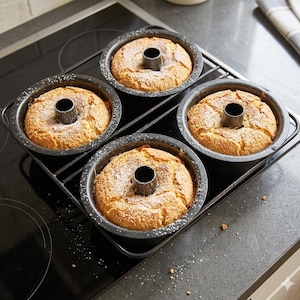 May include: Four freshly baked bundt cakes in individual round pans, dusted with powdered sugar, sit on a black metal cooling rack. The cakes have a golden-brown crust and a hole in the center. The rack is on a black stovetop.
