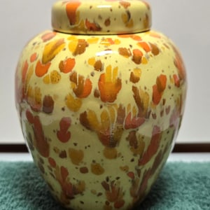 May include: A ceramic ginger jar with a lid, featuring a glossy pale yellow base color decorated with an abstract splatter pattern in shades of orange, brown, and dark yellow. The jar has a rounded body and a matching removable lid on top.