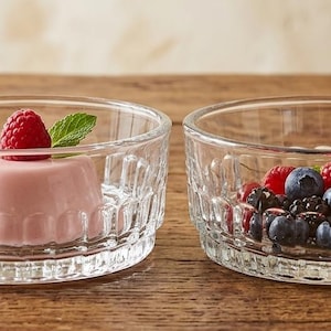 May include: Two clear glass dessert bowls on a wooden table. One bowl contains a pink dessert topped with a raspberry and mint leaf. The other bowl is filled with blueberries and raspberries. A spoon and rolled napkin are in the background.