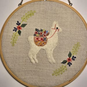 May include: Embroidered wall art featuring a white llama carrying a basket of colorful flowers. The llama is centered on a linen-textured background, framed by a wooden hoop. Floral accents in green, red, and blue adorn the border.