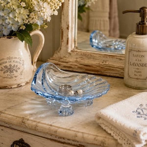 May include: A light blue glass dish shaped like a seashell with three small feet. The dish holds a silver ring and a small pearl earring. It is positioned near a cream-colored ceramic pitcher, a soap dispenser labeled Savon a la Lavande, and a towel.