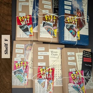 May include: Five rectangular books wrapped in brown and blue paper, tied with white string. Each features a 'Fizz Wiz' candy packet, a bookmark, and small paper labels with text like 'chilling', 'survival', 'romantasy', and 'post apocalyptic'.