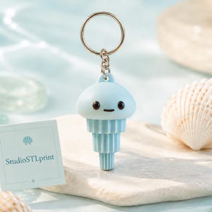 May include: A light blue jellyfish-shaped keychain with a silver keyring. The keychain has a smiling face and a tiered body. A small card with the text "StudioSTLprint" is also visible.