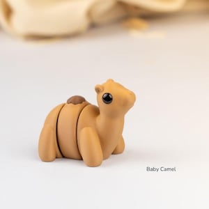 May include: A tan-colored baby camel figurine with a small brown hump and black eyes. The figurine is on a white surface with a soft, blurred background. The text "Baby Camel" is in the lower right corner.