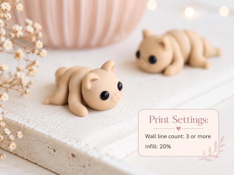 May include: Two small, tan-colored, handmade cat figurines with black eyes are lying on a white surface. The figurines are in a relaxed pose. The image also includes a blurred pink bowl and text that reads "Print Settings: Wall line count: 3 or more, Infill: 20%."