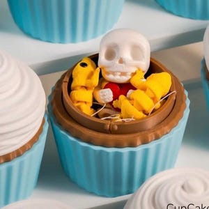 May include: A close-up of several cupcakes with blue liners and white frosting. One cupcake has a brown container with a white skull and yellow and red decorations. The text "CupCake" is visible in the bottom right corner.