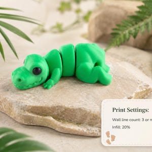 May include: A bright green articulated dinosaur toy with black eyes, resting on a light-colored stone. The toy is segmented, allowing for movement. The image includes a small card with print settings.