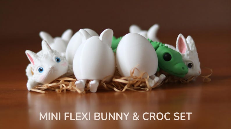 May include: A collection of small white bunny figurines and a green crocodile figurine, some emerging from white egg shells. The set is arranged in a cluster of straw-like material. The text MINI FLEXI BUNNY & CROC SET is printed below the figures.