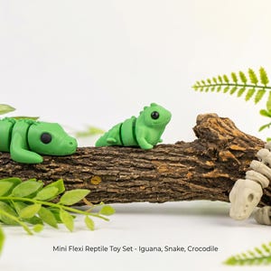 May include: A set of three articulated reptile toys including a green crocodile, a green iguana, and a beige snake. The figures are positioned on a piece of textured brown wood. The text "Mini Flexi Reptile Toy Set - Iguana, Snake, Crocodile" is printed below the figures.