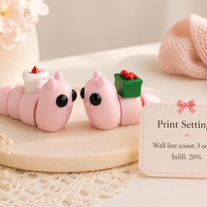 May include: Two pink caterpillar figurines with black eyes and small accessories. One has a white cake with a red candle, the other a green gift box with red bows. A sign reads "Print Settings: Wall line count: 3 or more, Infill: 20%."