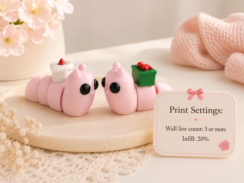 May include: Two pink caterpillar figurines with black eyes and small accessories. One has a white cake with a red candle, the other a green gift box with red bows. A sign reads "Print Settings: Wall line count: 3 or more, Infill: 20%."