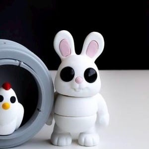 May include: A white bunny figurine with large black eyes, pink inner ears, and a small pink nose. Beside the bunny is a gray circular object with a small white chicken figurine inside. The background is black and white.