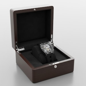 May include: A luxury watch displayed in a wooden watch box. The watch has a black strap and a rectangular face with visible internal mechanisms. The watch box is dark brown with a white top and a black velvet interior.