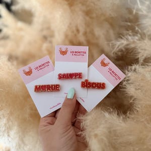 Peut inclure: Trois barrettes à cheveux rouges à paillettes sur des supports en carton blanc, affichant les mots MORUE, SALOPE et BISOUS. Les supports portent le texte LES BICHETTES À PAILLETTES avec une icône de cerf et des cœurs rouges intégrés.