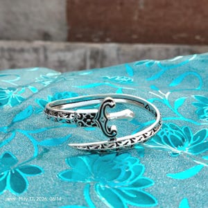 Op de afbeelding: Een zilverkleurige armband met een open ontwerp, met een zwaardvormig middelpunt en decoratieve patronen. De armband ligt op een turquoise stof met een bloemenpatroon. De afbeelding bevat de tekst "janni May 17, 2026, 06:14".