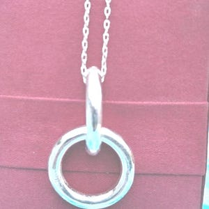 Può includere: Una collana color argento con una catenina sottile e un ciondolo composto da due anelli circolari lisci intrecciati. Il metallo ha una finitura lucida e riflettente che cattura la luce, creando un design pulito e semplice per il gioiello.