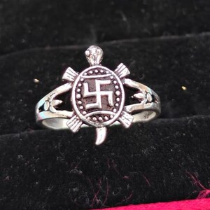 May include: A silver-toned metal ring featuring a turtle-shaped centerpiece. The turtle's shell is circular and displays a central geometric swastika symbol surrounded by a beaded border. The ring band splits into two arms on each side of the turtle.