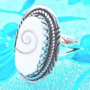 Op de afbeelding: Een zilveren ring met een ovaalvormige inleg van een witte shiva-oogschelp. De metalen zetting heeft een decoratieve touwachtige rand en een gekartelde rand. De ringband is gesplitst in twee aparte zilveren schachten die naar het middenstuk lopen.