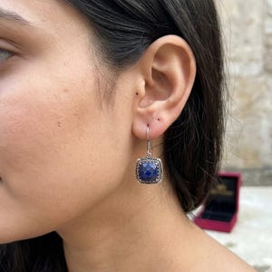 May include: A silver drop earring featuring a square, faceted deep blue gemstone set in a textured, oxidized silver frame with a rope-like border, suspended from a simple silver hook wire that passes through the earlobe.
