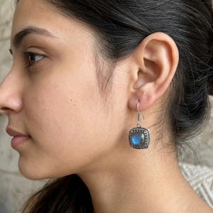May include: A silver drop earring featuring a square-shaped labradorite gemstone with a blue iridescent sheen. The stone is set in a decorative silver frame with a textured border and hangs from a simple silver hook wire finding.