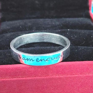 Peut inclure: Une bague en métal argenté avec l'inscription gravée "I am enough" en écriture cursive. La bague est placée à l'intérieur d'une boîte à bijoux rouge avec une doublure en velours noir, mettant en valeur la finition métallique et le texte.