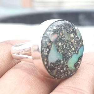 May include: A silver ring with a large, oval-shaped stone. The stone has a dark gray base with flecks of white and patches of light blue and green. The ring band is a simple, split design.