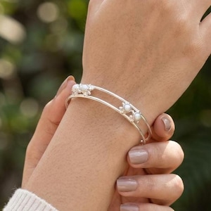 Puede incluir: Una pulsera tipo brazalete de plata con dos bandas paralelas conectadas por pequeños elementos decorativos. Cada elemento está engastado con una pequeña cuenta redonda de color blanco similar a una perla. Tiene un acabado metálico pulido.