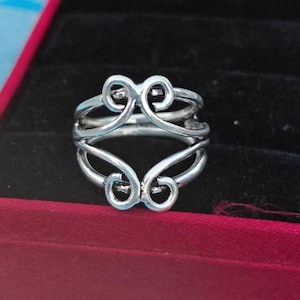 May include: A silver-toned metal ring featuring an openwork design with two symmetrical, swirling heart-like motifs positioned at the top and bottom of the band, creating a continuous, interconnected pattern around the circumference of the ring.