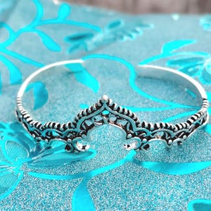 Puede incluir: Una pulsera plateada con forma de corona con intrincados detalles de filigrana. La pulsera tiene una banda curva y una pieza central decorativa con un diseño simétrico. Sobre una tela azul claro con un estampado floral.