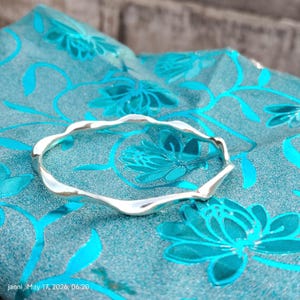 May include: A silver-coloured, wavy bangle bracelet. The bracelet is displayed on a teal fabric with a floral pattern. The fabric has a sheen, and the bracelet reflects light. The image includes the text "janni May 17, 2026, 06:20".