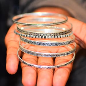 Op de afbeelding: Een stapel van zes zilverkleurige metalen armbanden, elk met een eigen reliëfpatroon. De ontwerpen omvatten gladde banden, ingewikkelde bloemmotieven, herhalende hartvormen en geometrische texturen, allemaal verticaal op elkaar gestapeld.