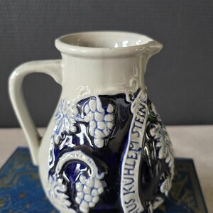 Könnte beinhalten: Ein weiß-blauer Keramikkrug mit Henkel und Ausguss. Der Krug hat ein Traubendesign und den Text "AUS KUHLEM STEIN". Der Krug steht auf einer blauen Oberfläche.