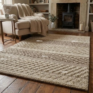 May include: A beige rectangular area rug with a textured, geometric pattern. The rug is placed on a hardwood floor in a living room setting, with a cozy armchair and fireplace in the background.